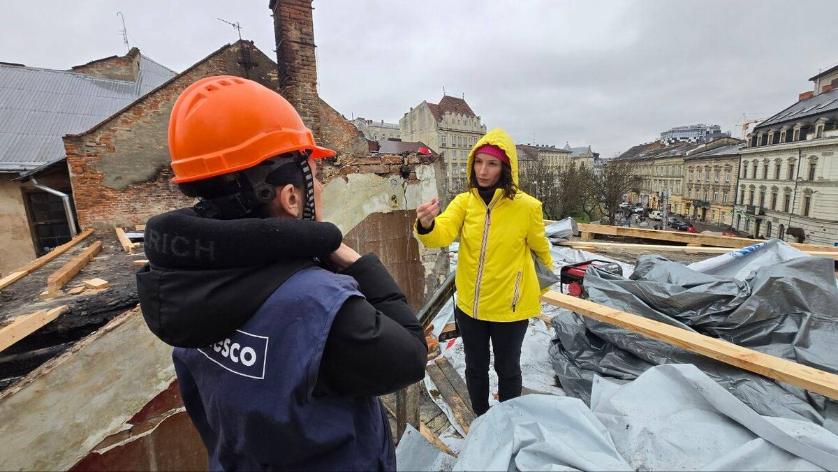 На пошкодженому даху будинку-пам'ятки на пл. Соборній, серед дерев&rsquo;яних балок, стоять обличчям одна до одної керівниця офісу охорони культурної спадщини ЛМР Тетяна Балукова у яскравій жовтій куртці та голова сектору культури Представництва ЮНЕСКО в Україні Марія Ріта Ачетозо, у синьому жилеті з написом &laquo;UNESCO&raquo; та помаранчевій касці. На другому плані, за рудими цегляними димарями, відкривається панорама історичних будівель Львова під похмурим небом.