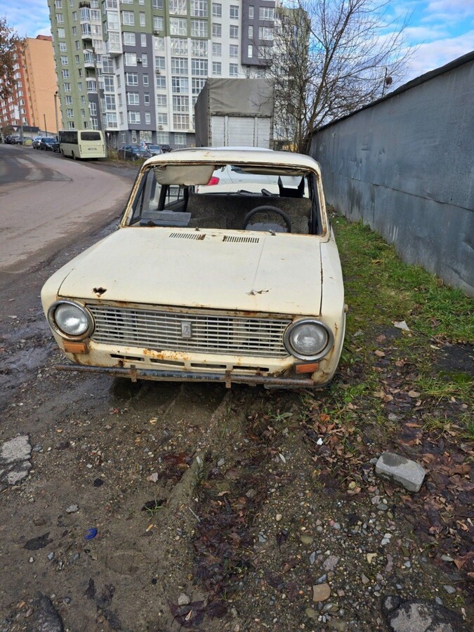 Покинутий, сильно поржавілий старий легковий автомобіль бежевого кольору стоїть на узбіччі. У машини розбите лобове скло, відсутні вікна. На другому плані видно сучасний багатоповерховий житловий будинок та пасажирський автобус і автівки