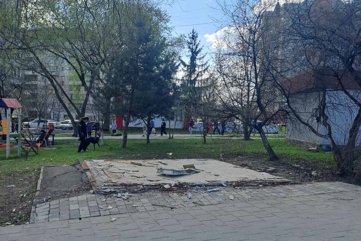 На землі біля тротуару видно залишки основи, на якій раніше стояв кіоск. Довкола ростуть дерева, збоку видно дитячий майданчик. Довкола гуляють люди з собаками