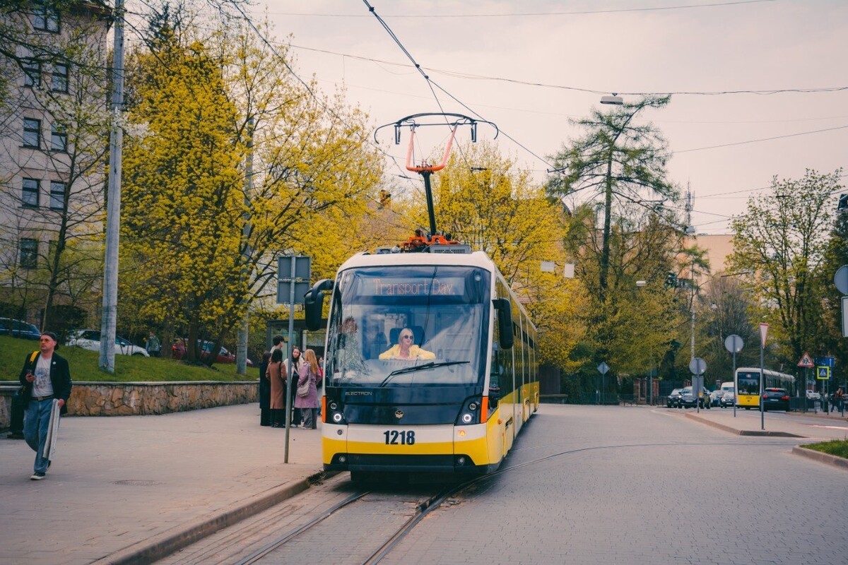 Трамвай &laquo;Електрон&raquo; з написом &laquo;Transport Day&raquo; на табло стоїть на рейках посеред вулиці,за кермом сидить водійка у жовтому одязі. Ліворуч на тротуарі біля зупинки стоїть група молодих людей, поруч проходить чоловік, а на другому плані видніються дерева та міські будівлі.