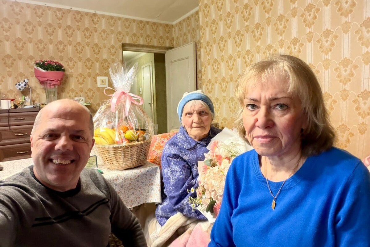 Столітня ювілярка Зінаїда Сидорова, її родичка та працівник управління соціального захисту Іван Фасольник сидять у кімнаті: ювілярка тримає в руках великий букет квітів, а поруч на столі стоїть «Кошик довголіття» зі смаколиками.