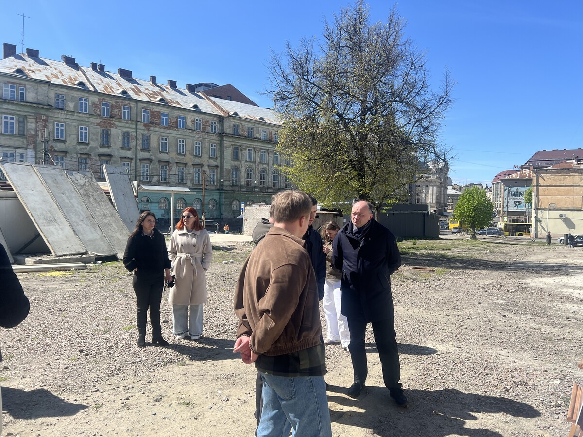 На фото посадовці і місцеві депутати під час виїзного огляду території колишнього ринку Добробут. Вони стоять на ділянці з гравійним покриттям. Поруч розташовані дерева та кілька тимчасових конструкцій, які демонтовують