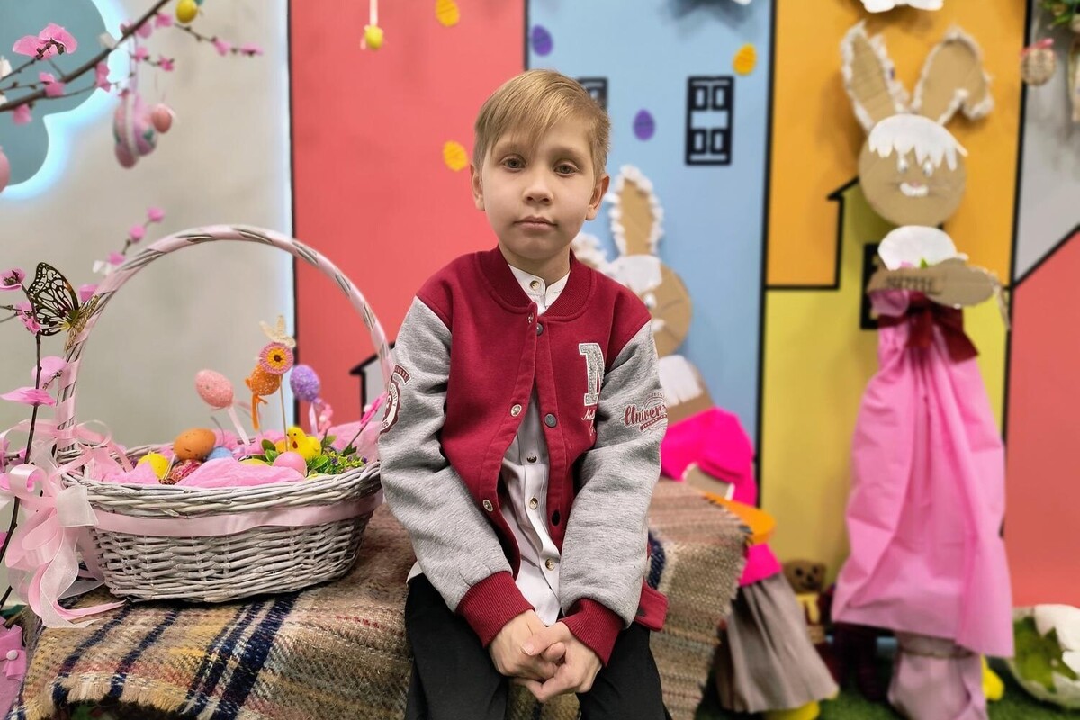 Пацієнт Юрчик сидить у на лавочці холі дитячої лікарні та спокійно і дещо замислено дивиться в камеру. Він одягнений у червоно-сіру куртку, а поруч із ним стоїть плетений кошик, наповнений декоративними яйцями та великодніми прикрасами.