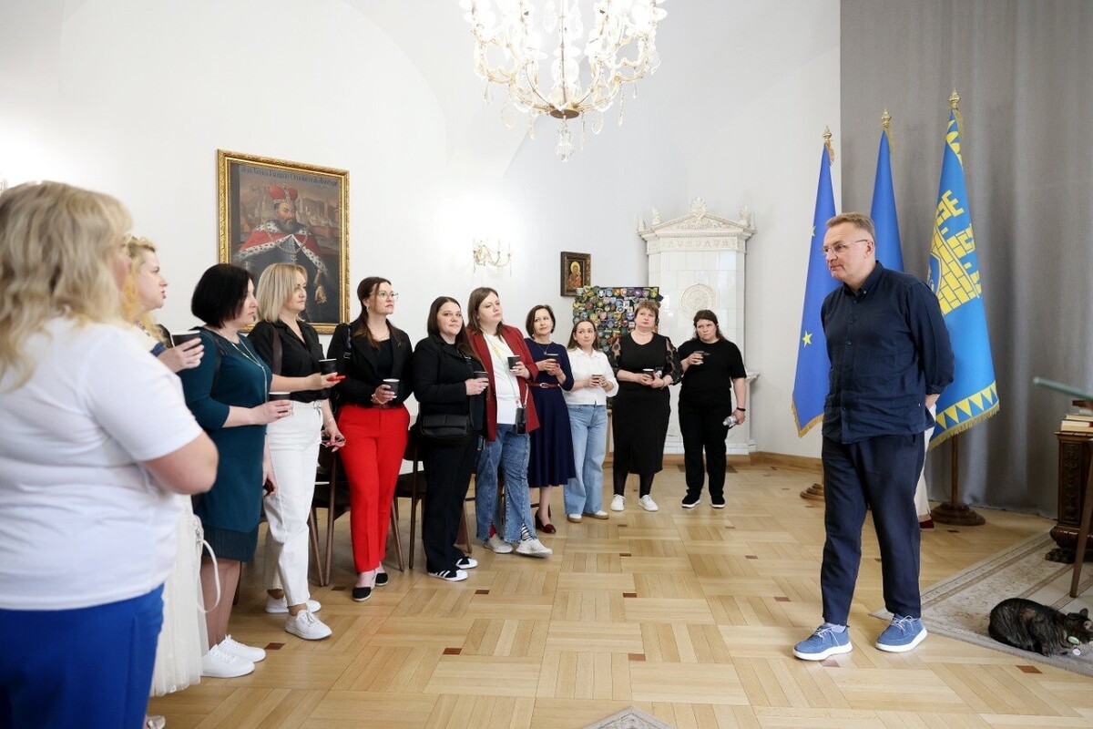 Мер Андрій Садовий дивиться на учасницю резиденції про соціальну політику Львова, яка розповідає про свої враження від навчальної програми. Вони знаходяться у кабінеті міського голови. На задньому плані видно сині прапори