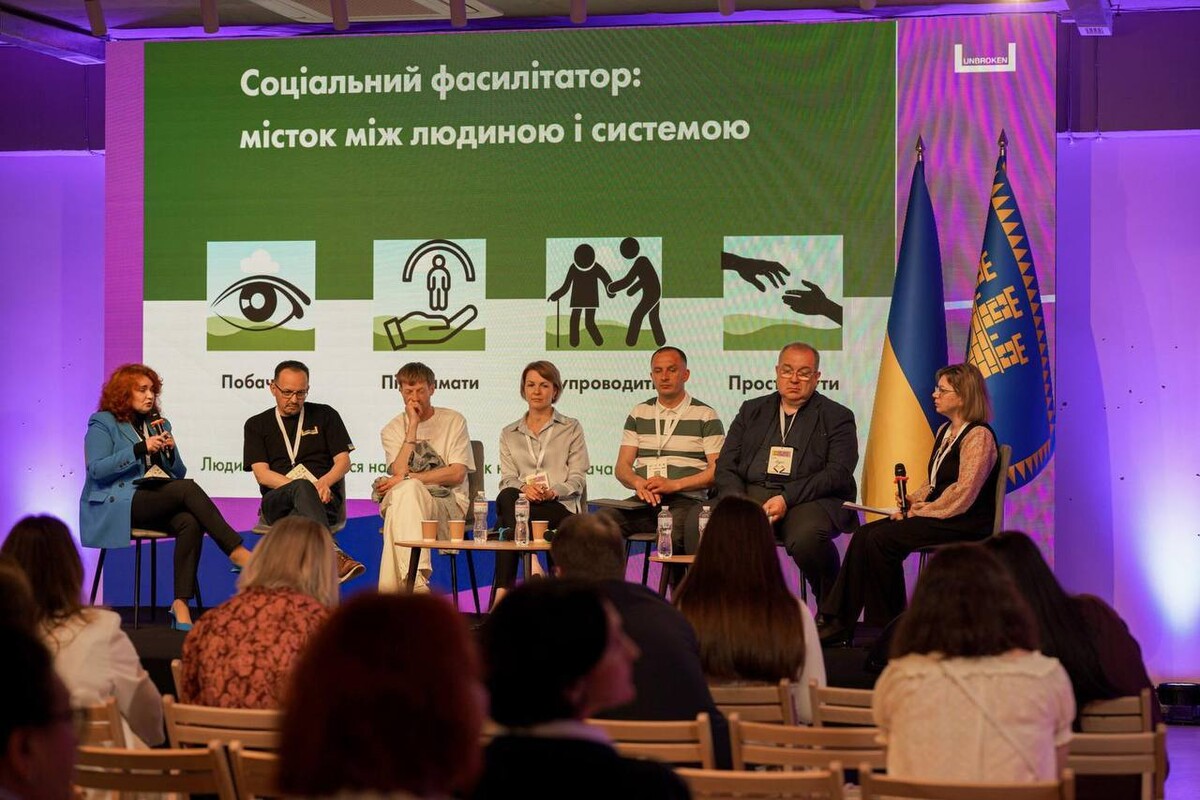 На сцені сидять спікери. Перед ними слухачі, які уважно слухають дискусію. На задньому фоні банер з написом "Соціальний фасилітатор: місток між людиною і системою"