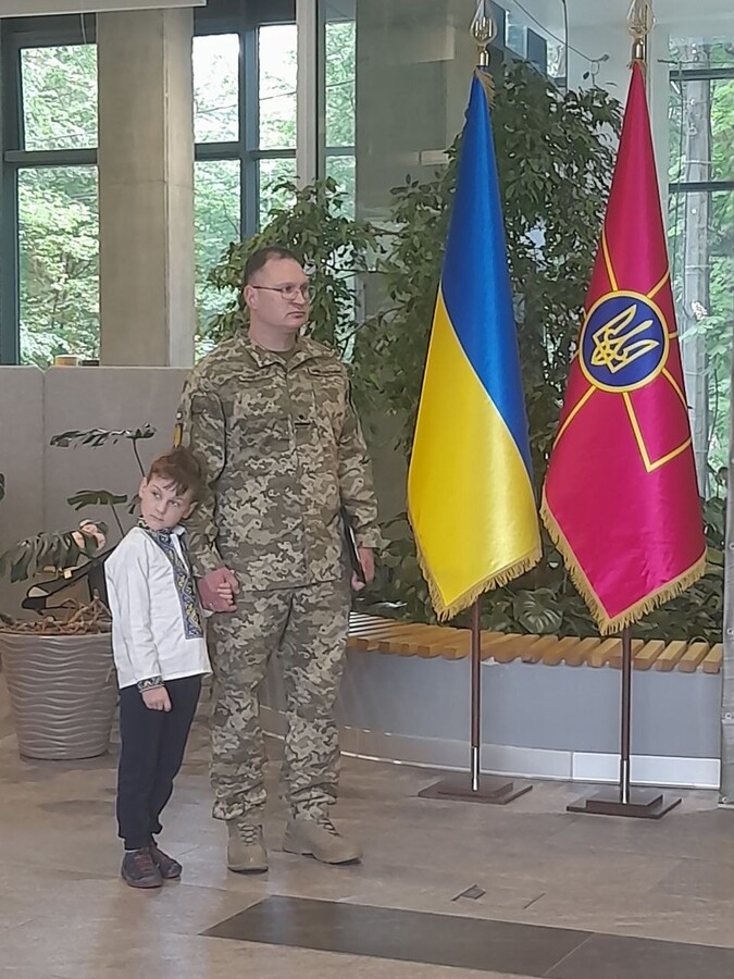 офіцер групи психологічної підготовки та супроводу управління психологічної підтримки персоналу ОК «Захід» майор Борис Кондратюк тримає за руку маленького хлопчика, який одягнений у вишиванцу. Вони стоять у приміщенні на тлі великих кімнатних рослин та двох прапорів: державного синьо-жовтого стяга України та малинового прапора ЗСУ із золотим тризубом.