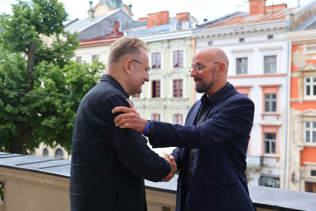 Міський голова Львова Андрій Садовий тисне руку Девіду Фортьє, засновнику фонду One World Strong Foundation на балконі львівської Ратуші. Довкола видно фасади старих історичних будинків центру міста. Збоку видно частину зеленої липи, що росте на площі Ринок
