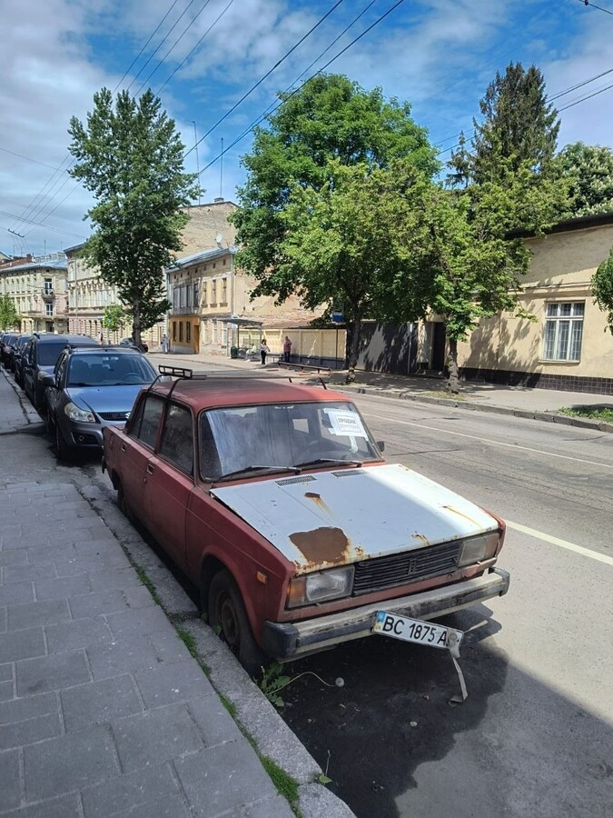 Припаркований на узбіччі вулиці старий червоний автомобіль з білим поіржавілим капотом, його передній номерний знак наполовину відірваний і тримається на одному кріпленні. На другому плані видо зелені дерева, будинки та інші автівки