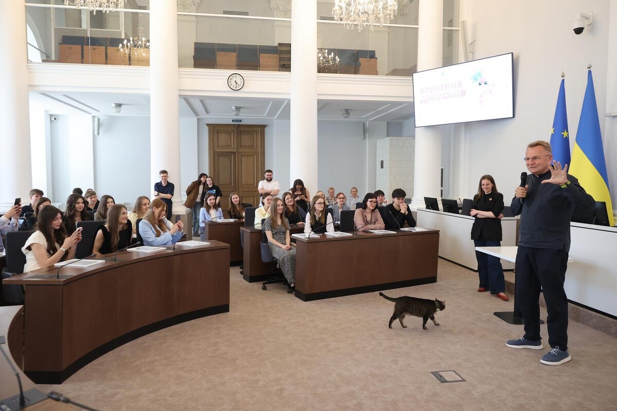 Випускники програми уважно слухають міського голову, який виступає перед ними. По центру зали ходить котячий мер Львова Левчик