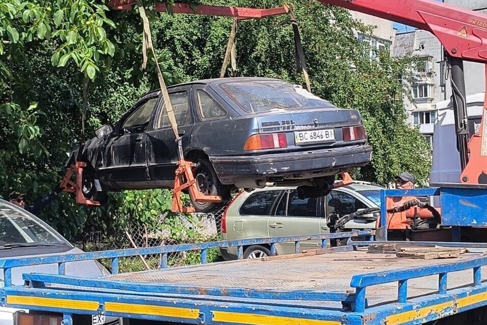 Процес евакуації покинутого автомобіля, який піднято в повітря за допомогою червоного крана-маніпулятора та спеціальних вантажних ременів, закріплених за колеса. Машину переміщують на платформу евакуатора, що стоїть на передньому плані на тлі зелених дерев та житлового будинку.