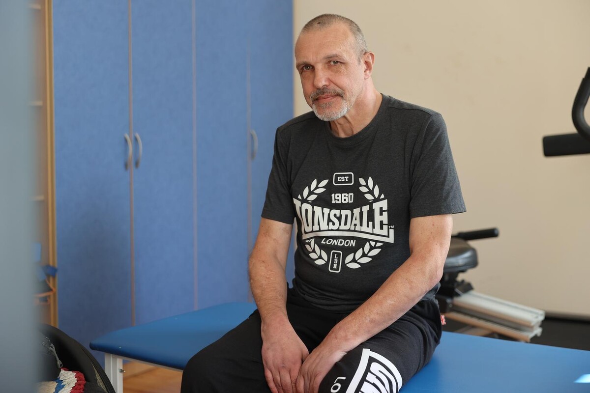 Юрій Альошин сидить на кушетці в реабілітаційному залі. Він дивиться прямо в камеру, погляд у нього замислений. Позаду видно сині шафки.