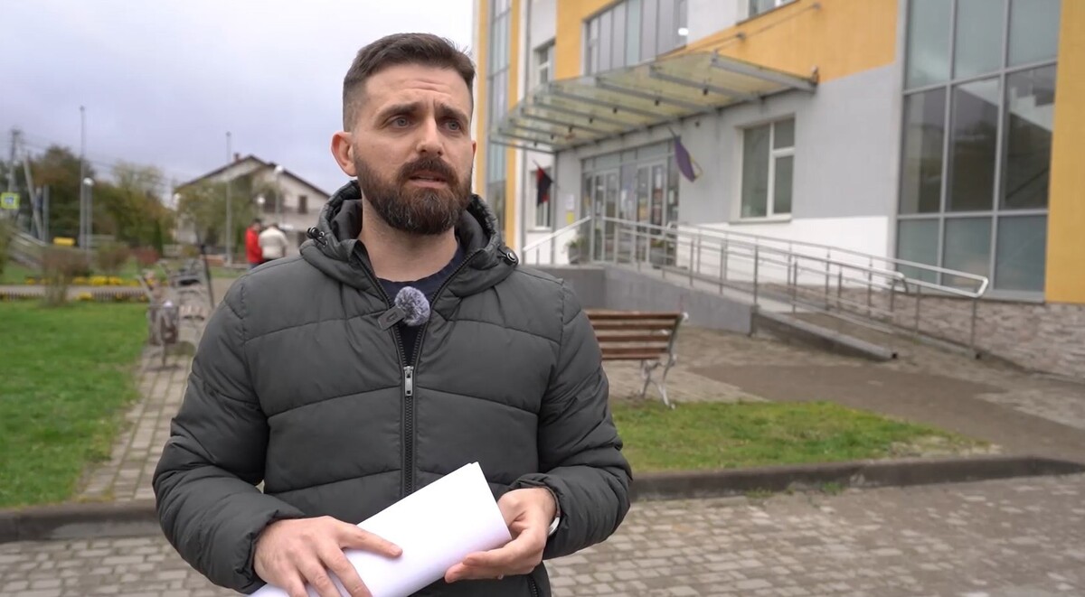 Чоловік стоїть поруч з будівлею народного дому в Сокільниках. В руках тримає документи. Позаду нього видно лавки, забруковану площу та перехожих.