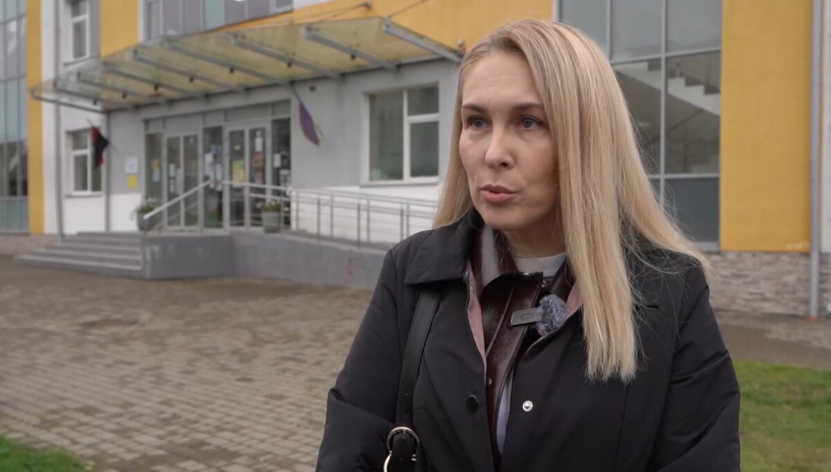 Жінка в чорному пальто стоїть перед будівлею народного дому в Сокільниках. Позаду неї видно головний вхід до будівлі та забруковану площу попереду