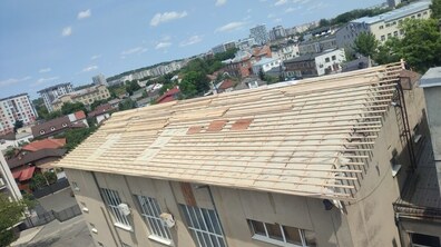 Фото зроблене з висоти - видно велику будівлю з двосхилим дахом, який перебуває в процесі ремонту. На даху змонтовані дерев'яні рейки та постелено мембрану. Навколо розташований міський район із приватними будинками, багатоповерхівками та великою кількістю дерев