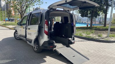 Спеціалізований автомобіль світло-сірого кольору, призначений для перевезення людей, які користуються кріслами колісними. Машина стоїть на брукованому майданчику просто неба в сонячний день.