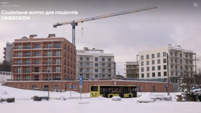 Три шестиповерхові будинки на вул. Миколайчука у Львові. В одному з них вже мешкають пацієнти, що проходять реабілітацію в центрі НЕЗЛАМНІ. Інші два ще добудовують. На майданчику видно будівельний кран. Довкола земля вкрита снігом. Дорогою їде великий автобус