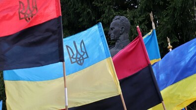 Жовто-блакитні та червоно-чорні стяги з Гербами України майорять на фоні пам’ятника Роману Шухевичу. На фоні гілля зеленої ялини