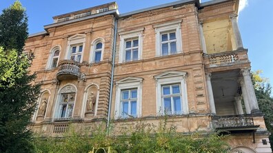 Старовинна двоповерхова будівля на вулиці Парковій з персиковими стінами та білими вікнами. Фасад прикрашений декоративними елементами, колонами, балконами та ліпниною. Навколо будинку ростуть дерева та кущі.