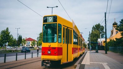 Жовто-червоний трамвай №9 стоїть на рейках біля зупинки. Праворучі видно церкву, ліворуч — дорогу з машинами та будівлю. З обох боків ростуть зелені дерева.