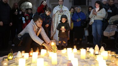Жінка схилилася над білою тканиною, розстеленою на землі, та ставить серед інших запалених свічок і лампадок скляний свічник із вогником. Поруч із нею стоїть маленький хлопчик із палаючою свічкою в руках, а позаду них зібралися інші люди та священник.