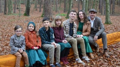 Яна Король із чоловіком та п’ятьма дітьми серед лісу сидять в один ряд на поваленому стовбурі дерева, вкритому помаранчевою тканиною.