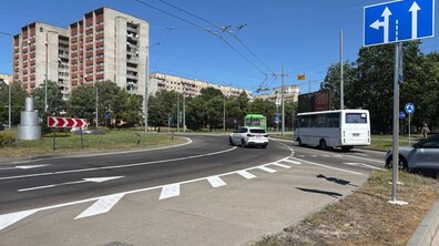 Міське перехрестя з кільцевим рухом, де на асфальті нанесено білу розмітку зі стрілками напрямку руху. У кадрі видно білий та зелений автобуси і білий легковий автомобіль, а на правому передньому плані - синій дорожній знак «Напрямки руху по смугах». На другому плані височіють багатоповерхові житлові будинки, також видно зелені дерева під ясним синім небом.
