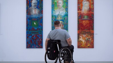 Чоловік на кріслі колісному розглядає три великі вертикальні картини, розміщені на білій стіні виставкової зали.