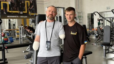Пацієнт – захисник Андрій Оленич та фізичний терапевт центру UNBROKEN стоять у реабілітаційному залі. Обоє дивляться в камеру й усміхаються. У захисника забинтовані кукси обох рук. Позаду видно різне обладнання для занять.