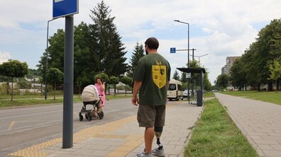 Чоловік із протезом ноги йде тротуаром із тактильною плиткою. Поруч дорога, де жінка переходить із дитячим візком. Видно зупинку транспорту, білий мікроавтобус і зелені дерева навколо.