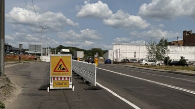Дорога, на якій встановлено тимчасове біле пластикове загородження з жовтим попереджувальним знаком «Дорожні роботи» та написом «УВАГА!». На задньому плані видно легкові автомобілі, вантажівки, промислові будівлі та зелені дерева.