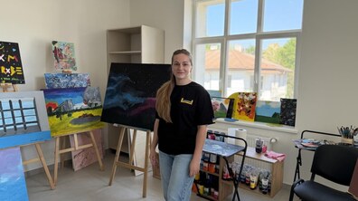 Усміхнена військова медикиня Марина Сергієнкова стоїть у світлій творчій майстерні центру ментального здоров’я святого Лева Великого у Львові. Навколо неї – багато картин на мольбертах, видно фарби та художні матеріали. Жінка одягнена в чорну футболку з лого UNBROKEN та світлі джинси.