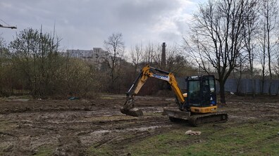 Невеликий екскаватор на болотистій ділянці на території парку Архангела Михаїла. Довкола дерева та кущі. Позаду здалеку видніються багатоповерхівки та інша забудова