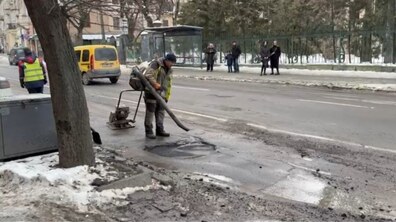 Працівник шляхово-ремонтного підприємства Личаківського району готує яму на дорозі до закладання холодним асфальтом. Зокрема, за допомогою ручної промислової повітродувки він здуває воду та бруд. Позаду працівника на дорозі стоїть ще один працівник та розміщена спеціальна ручна установка для розгладження засипаного асфальту. З протилежного боку вулиці видно зупинку громадського транспорту та людей, які очікують на транспорт. Дорогою проїжджає автомобіль жовтого кольору.