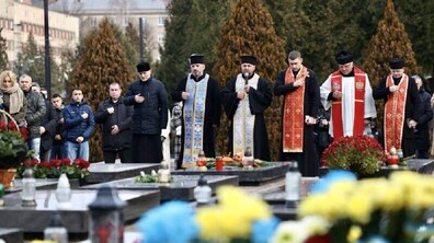 На фото — урочиста поминальна церемонія на військовому цвинтарі. Попереду видно ряди могил зі квітами та лампадками. Біля могил - священнослужителі у рясах та кілька цивільних.