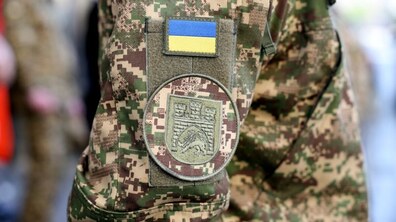 Крупним планом зображено рукав військової форми з камуфляжним візерунком. На ньому прикріплений шеврон із прапором України, а нижче є інший нашивний знак із символікою підрозділу. Фон розмитий, що підкреслює деталі форми.