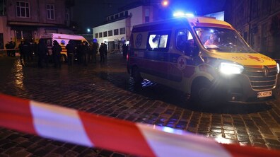 Місце, де пролунали вибухи. У центрі кадру стоїть автомобіль швидкої допомоги з увімкненими синіми проблисковими маячками. В салоні видно медика. На задньому плані — ще одна карета «швидкої», група людей та правоохоронців, які зібралися біля будівель.