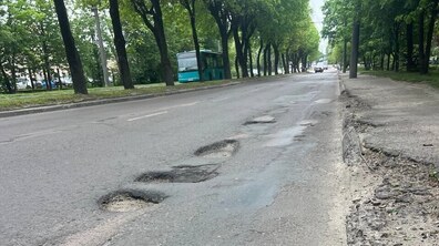 Вулиця Сяйво з алеєю високих дерев обабіч. Уздовж правого краю дороги видно кілька великих вибоїн і пошкоджений асфальт. Дорога майже порожня: вдалині їде кілька авто, а ліворуч стоїть зелений автобус.