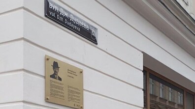 На світлому фасаді будинку видно чорну табличку з назвою вулиці — Джохара Дудаєва. Нижче встановлена пам’ятна металева таблиця з портретом Джохара Дудаєва та коротким текстом про нього. Праворуч — велике вікно, у якому віддзеркалюються будинки навпроти.
