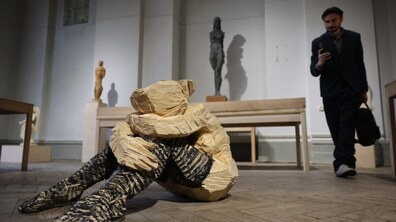 На фото зображено виставкову залу з сучасною дерев’яною скульптурою людини, що сидить, обійнявши коліна. На задньому плані видно інші фігури й відвідувача, який розглядає експозицію та знімає її на телефон.