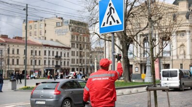Працівник у червоній формі фарбує стійку дорожнього знака «пішохідний перехід» в центрі Львова на фоні Оперного театру