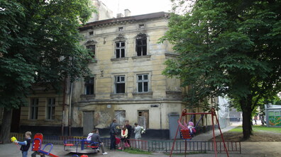 Триповерховий історичний будинок в аварійному стані без вікон, та з розбитими шибками. Перед ним дитячий майданчик, на якому бавляться діти