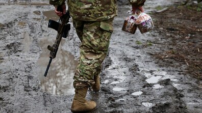 Військовий у формі йде по розбитій болотяній дорозі. В одній руці у нього паска, а в іншій автомат