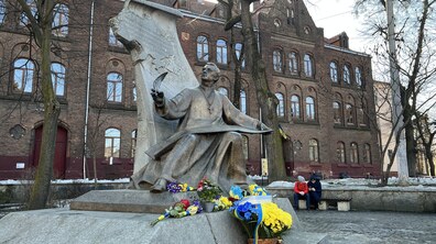 Бронзовий пам’ятник Михайлу Вербицькому, який сидить із розгорнутим сувоєм та пером у руці. Біля підніжжя пам’ятника квіти з синьо-жовтими стрічками та запалені лампадки. Позаду видно цегляну історичну будівлю — школу № 3, а на лавці праворуч сидять двоє людей