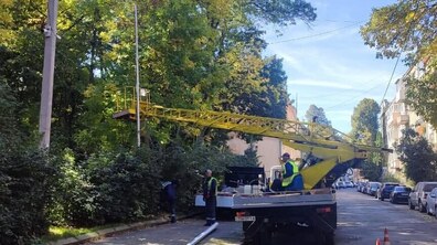 На міській вулиці працівники комунальних служб виконують роботи із заміни світильників з використанням автовишки. Неподалік також стоїть вантажівка. Біля неї працюють кілька комунальників у світловідбивних жилетах.