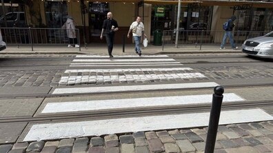 Міська вулиця з пішохідним переходом через бруківку й трамвайні колії. Двоє чоловіків переходять дорогу. На задньому плані — будівля з магазинами, кілька людей і припарковане авто