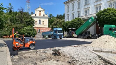 укладання асфальту на вул. Винниченка на фоні храму священномученика Климентія Шептицького УГКЦ. Ліворуч на свіжому чорному асфальті працює помаранчевий дорожній каток із водієм. По центру стоїть будівельна тачка на розритій ділянці, а праворуч насипана велика купа піску. Праворуч велика зелена вантажівка з піднятим кузовом та синій самоскид із причепом. Поруч працюють робітники.