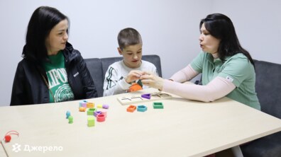 Дитина сидить за столом разом із двома соціальними працівницями: одна з них допомагає хлопчику тримати невеликий предмет, ймовірно пояснюючи або навчаючи його, а інша працівниця спостерігає за процесом із зацікавленням. На столі лежать різнокольорові геометричні фігури
