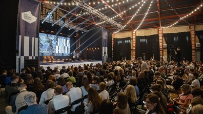 Фото із торішнього Lviv Urban Forum: велике просторе приміщення з індустріальним інтер'єром. У залі присутня велика кількість людей, які сидять на стільцях рядами та слухають виступ. На великому екрані напис — Lviv Urban Forum.