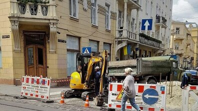 Ремонтні роботи на вулиці міста. У центрі — жовтий компактний екскаватор, за ним стоїть вантажівка-самоскид, а на передньому плані видно дорожні огородження та жінку-пішохода, яка проходить повз.