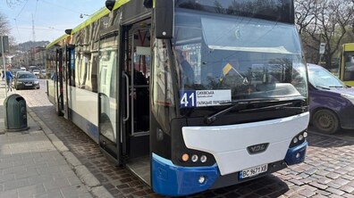 Сучасний великий низькопідлоговий міський автобус стоїть на зупинці на вузькій брукованій вулиці, двері відчинені для посадки пасажирів. На табло видно маршрут №41. Навколо — автомобілі, на другому плані видно будинки